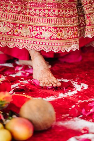 Bride's henna-decorated foot stepping on red rose petals beside a coconut and fruit during a traditional Indian wedding ritual.