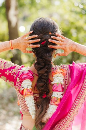 Bride seen from behind with a long braided hairstyle, henna-decorated hands raised, pink silk saree and floral garland in a sunlit garden.