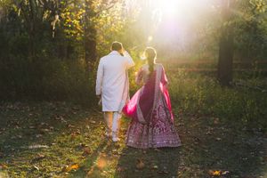 Couple in traditional Indian wedding attire walking hand in hand through a sunlit forest, with golden light streaming through the trees.