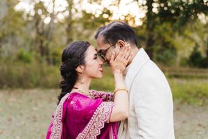 Couple sharing an intimate forehead touch outdoors, she wears a magenta embroidered sari and he wears a cream jacket amid autumn trees.