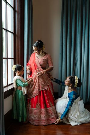 A bride in a red and gold embroidered lehenga is helped by a young girl and a kneeling woman beside a tall window with teal curtains.