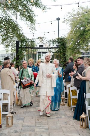 Groom in a cream sherwani and turban walks down an outdoor aisle with guests, a musician, and string lights at an Indian wedding.