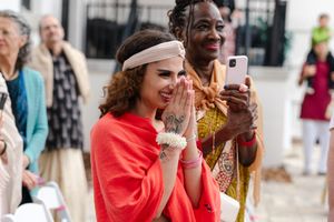 A smiling woman in an orange-red shawl and cream headband presses her hands together at an outdoor wedding as guests look on.