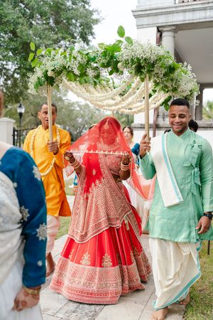 Indian bride in a red and gold lehenga walks under a floral canopy held by men in traditional attire during an outdoor wedding procession.