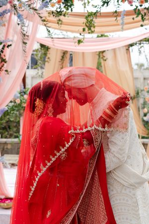 Bride in red embroidered lehenga and sheer veil embraces groom in ivory sherwani beneath a floral wedding canopy.