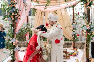 A bride in a red lehenga and a groom in a cream sherwani stand together beneath a floral wedding mandap draped in soft fabric.