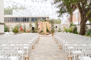 Outdoor wedding ceremony setup with white chairs lining a stone aisle leading to a floral arch with blush drapes under string lights.
