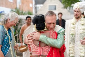 A man in a green kurta embraces a bride in a red and coral lehenga as guests in floral garlands look on at an Indian wedding.