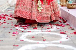 Bride in an ornate red and gold embroidered lehenga stands barefoot on stone tiles scattered with red rose petals and white rangoli designs.