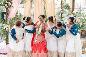 Bride in a red and gold sari laughs with four bridesmaids in white and blue saris at a floral-decorated outdoor Indian wedding.