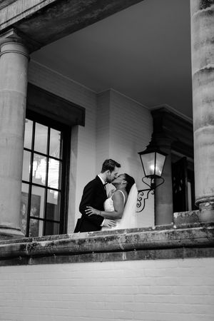 Black and white photo of a bride and groom sharing a kiss on a stone balcony beside a vintage lantern and tall columns.