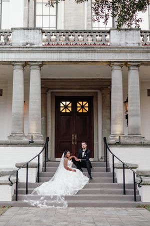 Bride in a flowing white gown and groom in a black tuxedo sit together on the steps of a grand columned building with wooden double doors.