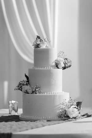 Black and white photo of a three-tier wedding cake with buttercream finish, pearl trim, and fresh rose and baby's breath florals.