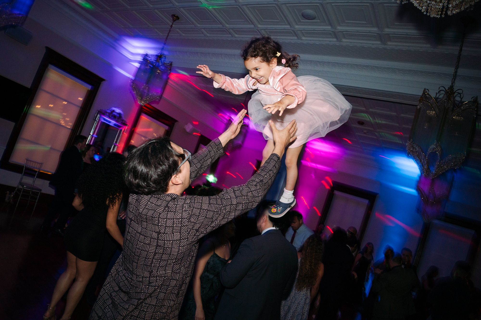Man in a plaid blazer tosses a laughing young girl in a pink dress into the air on a party dance floor lit with purple and blue lights.