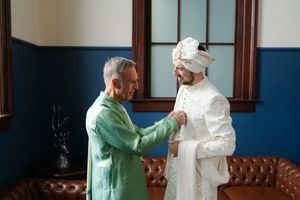 Older man in a green silk kurta adjusts the cream sherwani of a groom wearing a turban in a room with blue walls and a brown leather sofa.