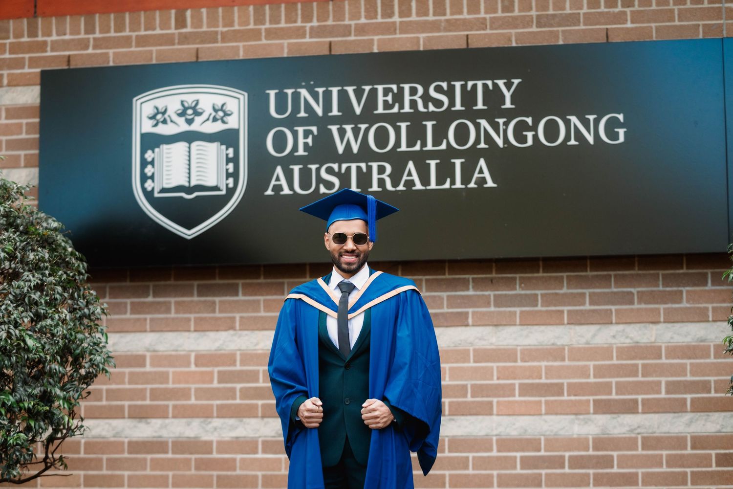 Smiling graduate in blue gown and cap poses in front of the University of Wollongong Australia sign on a brick wall.