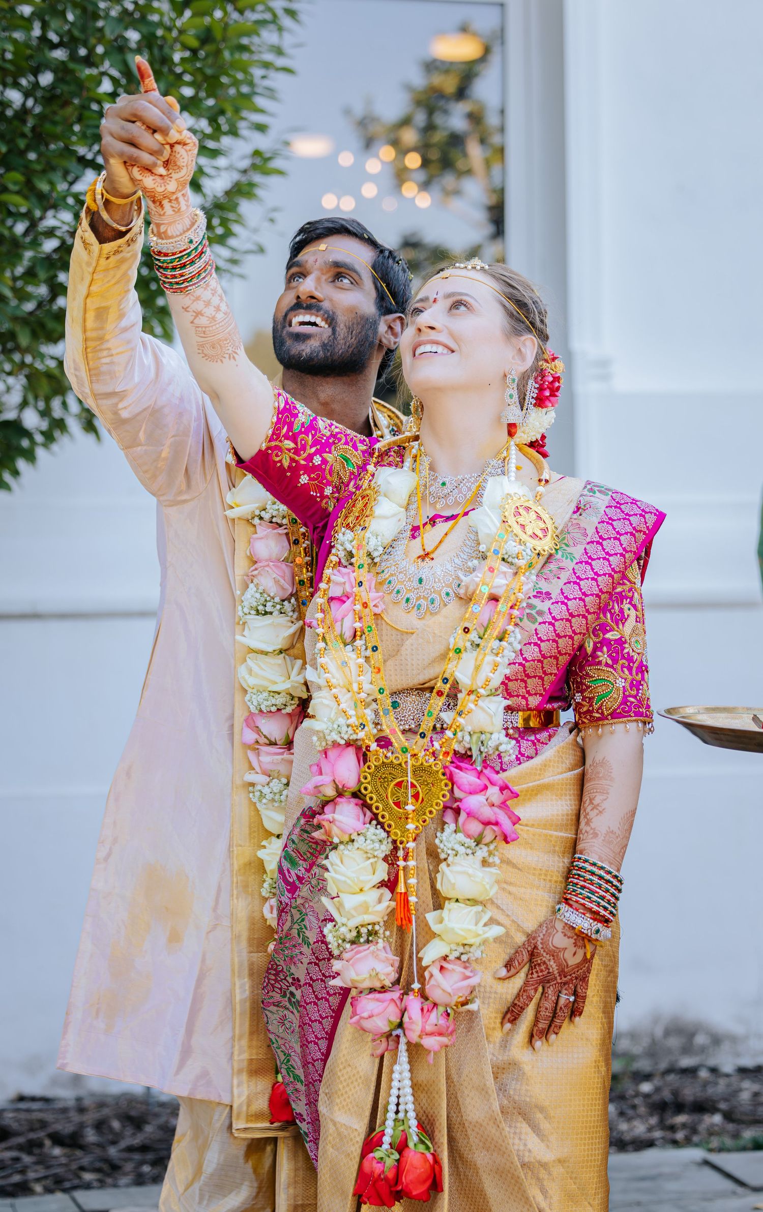 Smiling couple in traditional Indian wedding attire with floral garlands, silk outfits, and henna-decorated hands pointing upward outdoors.