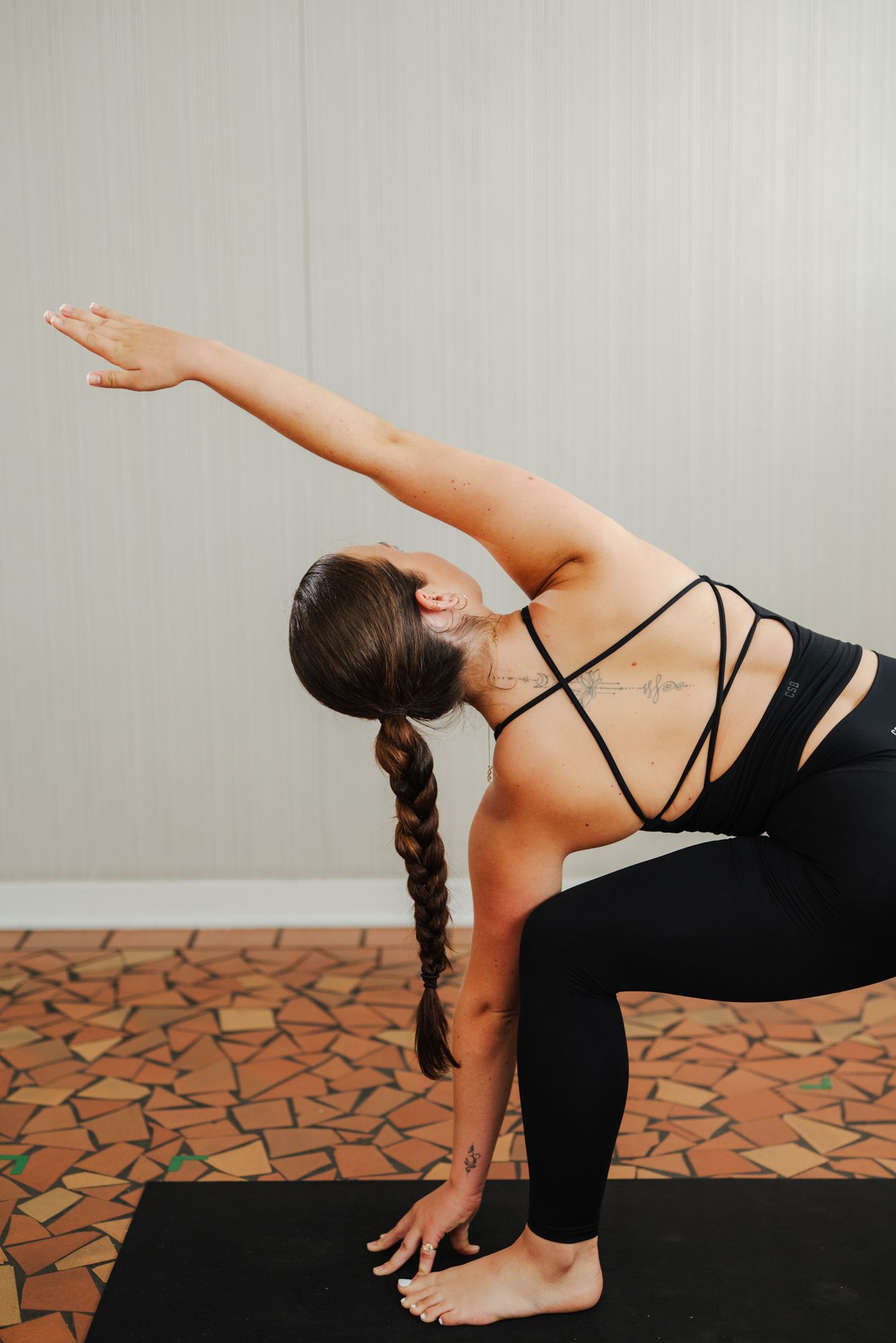 Woman with a long braid practices a revolved triangle yoga pose on a black mat, wearing black leggings and a strappy top on a tiled floor.