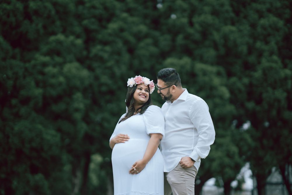 Expectant couple embracing outdoors during a maternity photoshoot, the mother wearing a white dress and flower crown beside green trees.