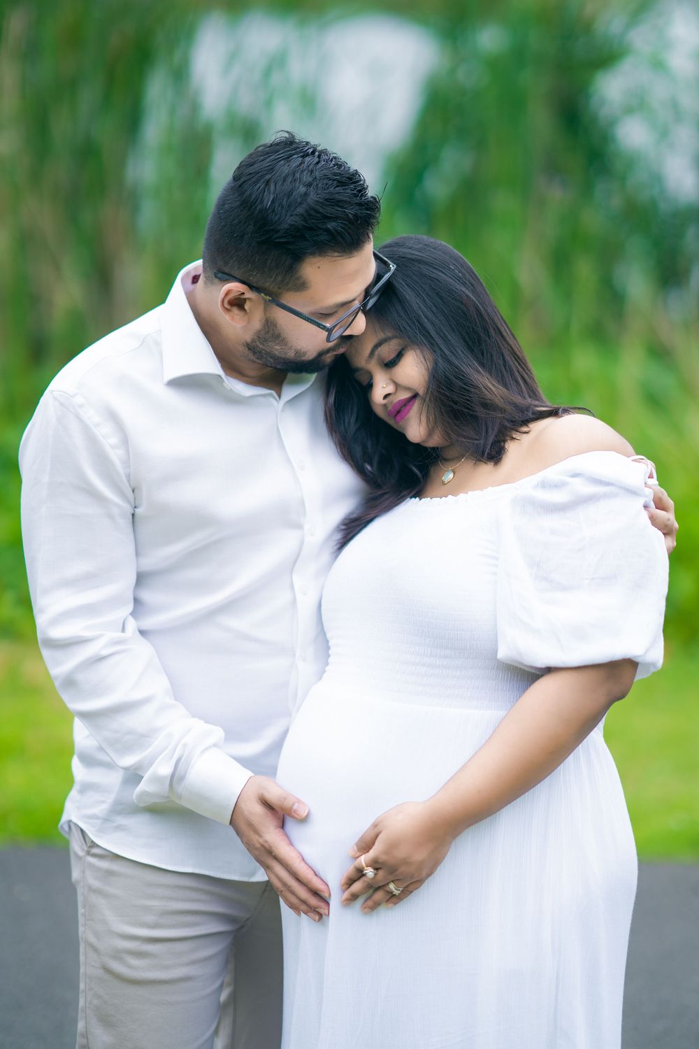 Expecting couple embracing outdoors, the man in a white shirt and glasses kissing his partner's forehead as she cradles her baby bump.