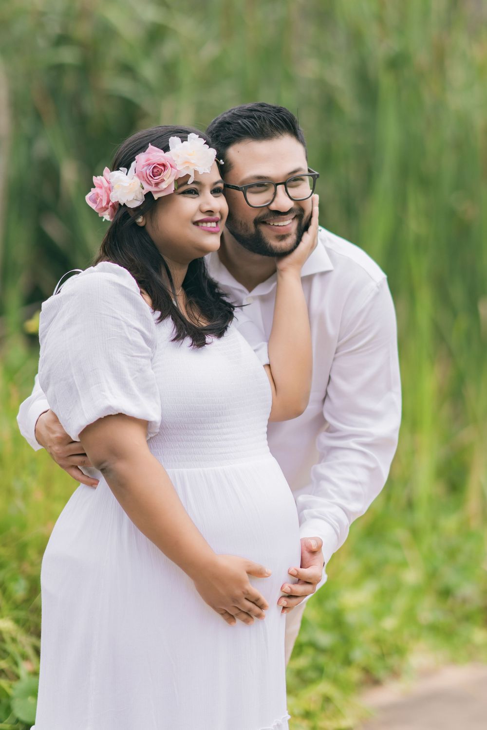 Smiling pregnant woman in a white dress and floral crown embraced by her bearded husband in glasses during an outdoor maternity photoshoot.