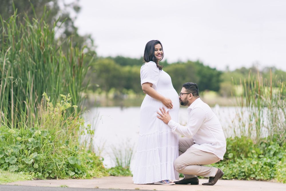 Smiling pregnant woman in a white dress stands by a lake as her kneeling partner gently cradles her baby bump in a maternity photoshoot.