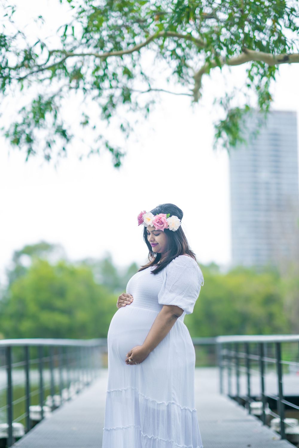 Pregnant woman in a white dress and floral flower crown cradles her baby bump on a wooden bridge with green trees and a city skyline behind.
