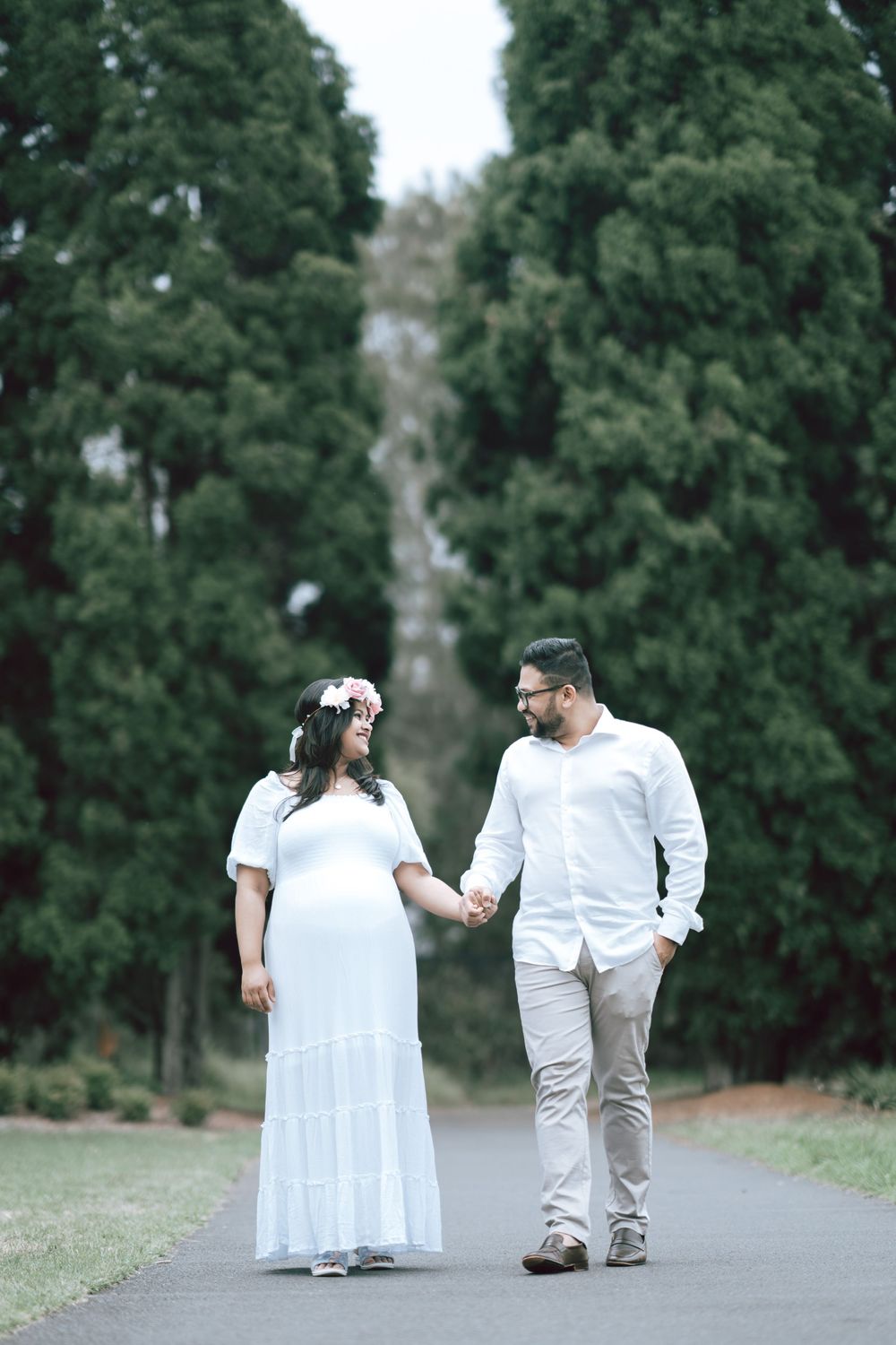 Couple holding hands and walking along a paved path lined with tall evergreen trees, she in a white dress with a floral crown.