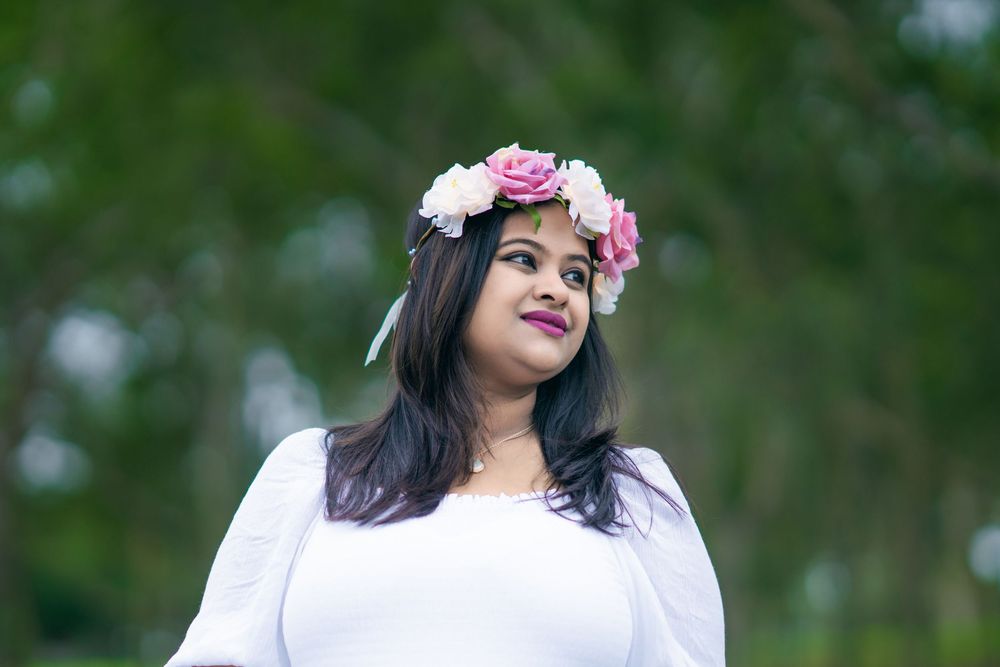 Smiling woman with long dark hair wearing a pink and white floral crown and a white dress, posing outdoors against a blurred green backgroun