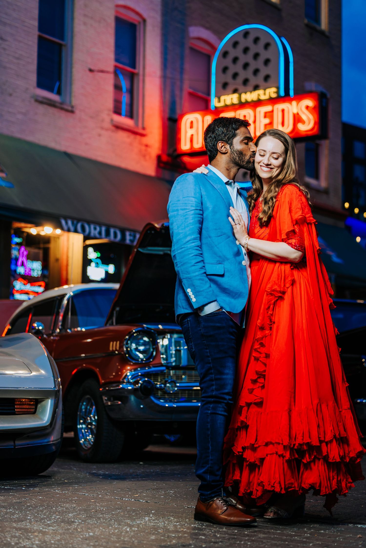 Couple in a blue blazer and flowing red ruffled gown share a kiss beside vintage cars under glowing neon signs on a city street at dusk.