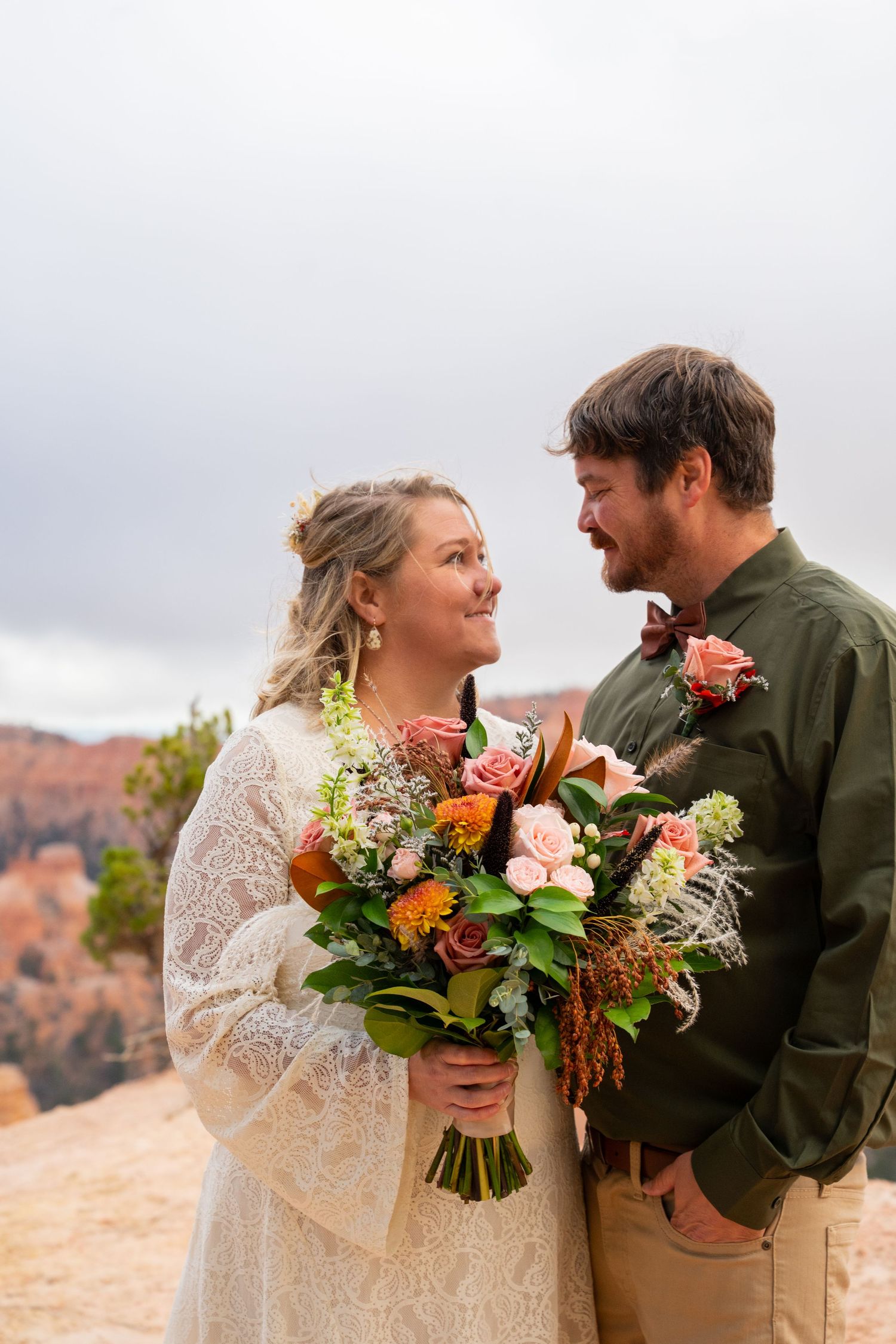 Bride in a lace dress and groom in an olive shirt smile at each other holding a colorful bouquet at a scenic desert canyon overlook.