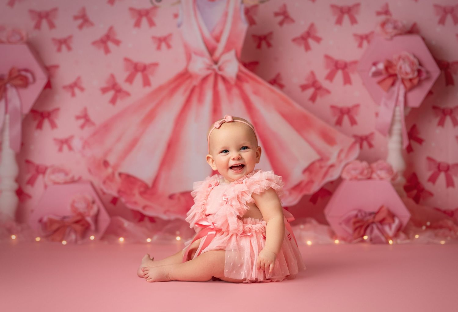 Baby wearing pink tutu dress sits on pink backdrop with bow pattern and ballerina dress decoration.