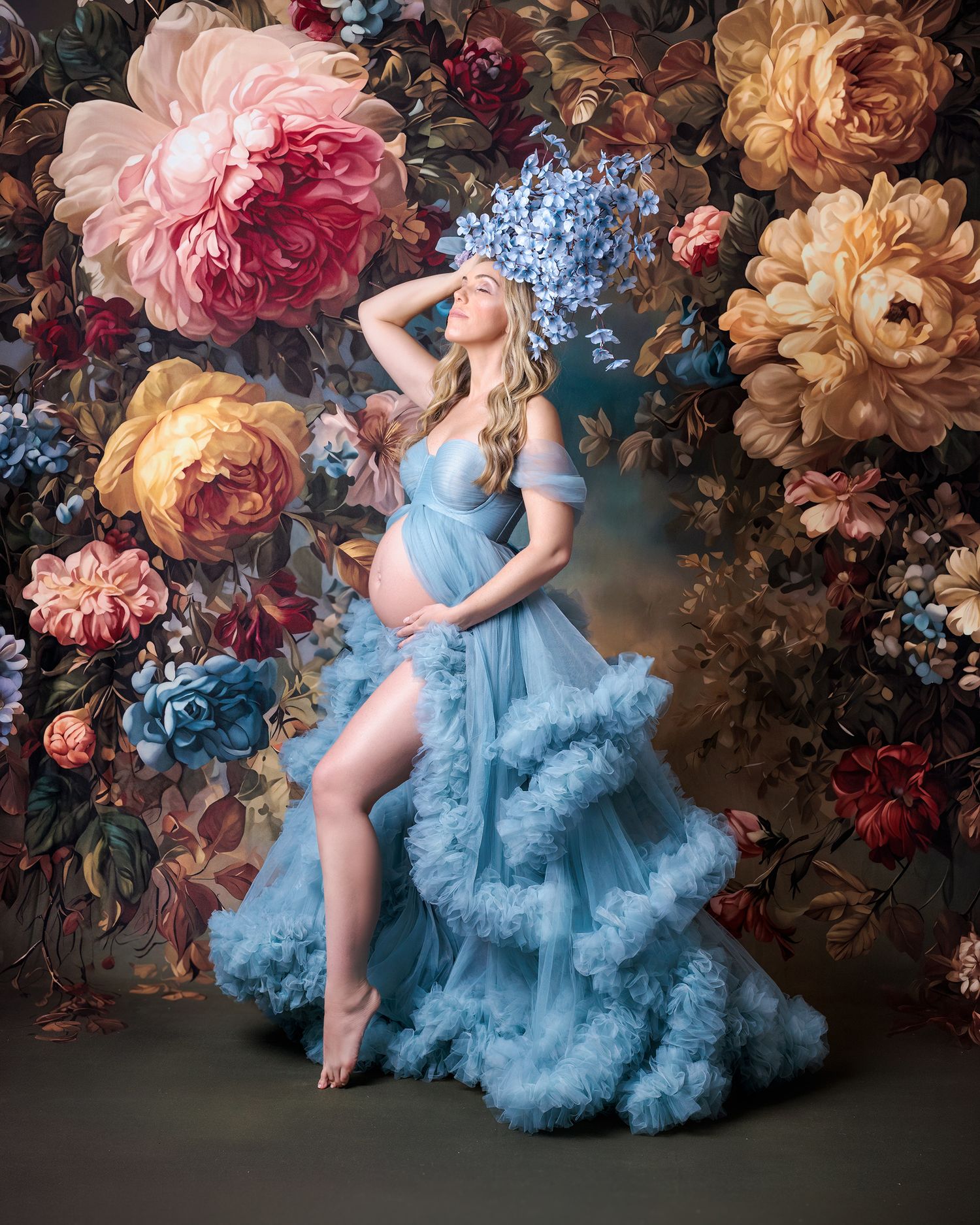 Pregnant woman stands against large bloom filled backdrop holding blue flower bouquet above hear head she lifts her face high to the light.