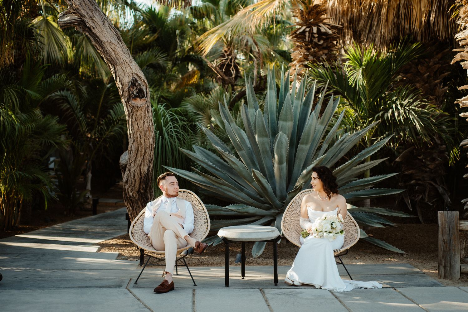 Elegant wedding couple seated in modern chairs amid tropical garden setting.