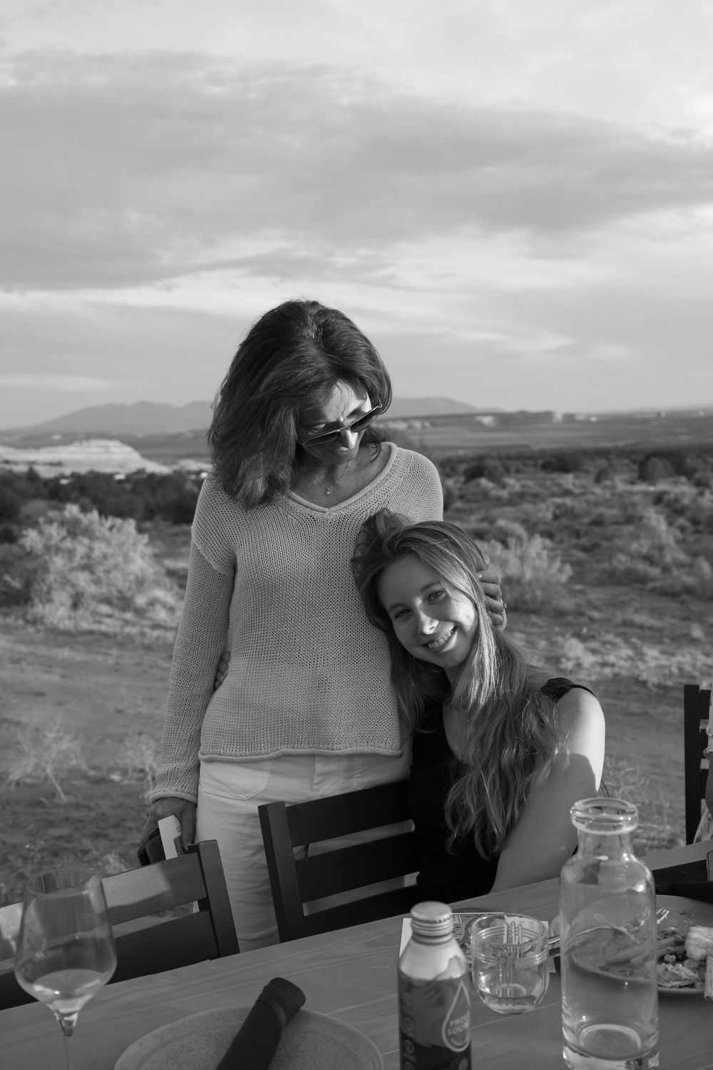 Black and white photo of two women sharing a warm moment at an outdoor dining table with scenic desert landscape backdrop.