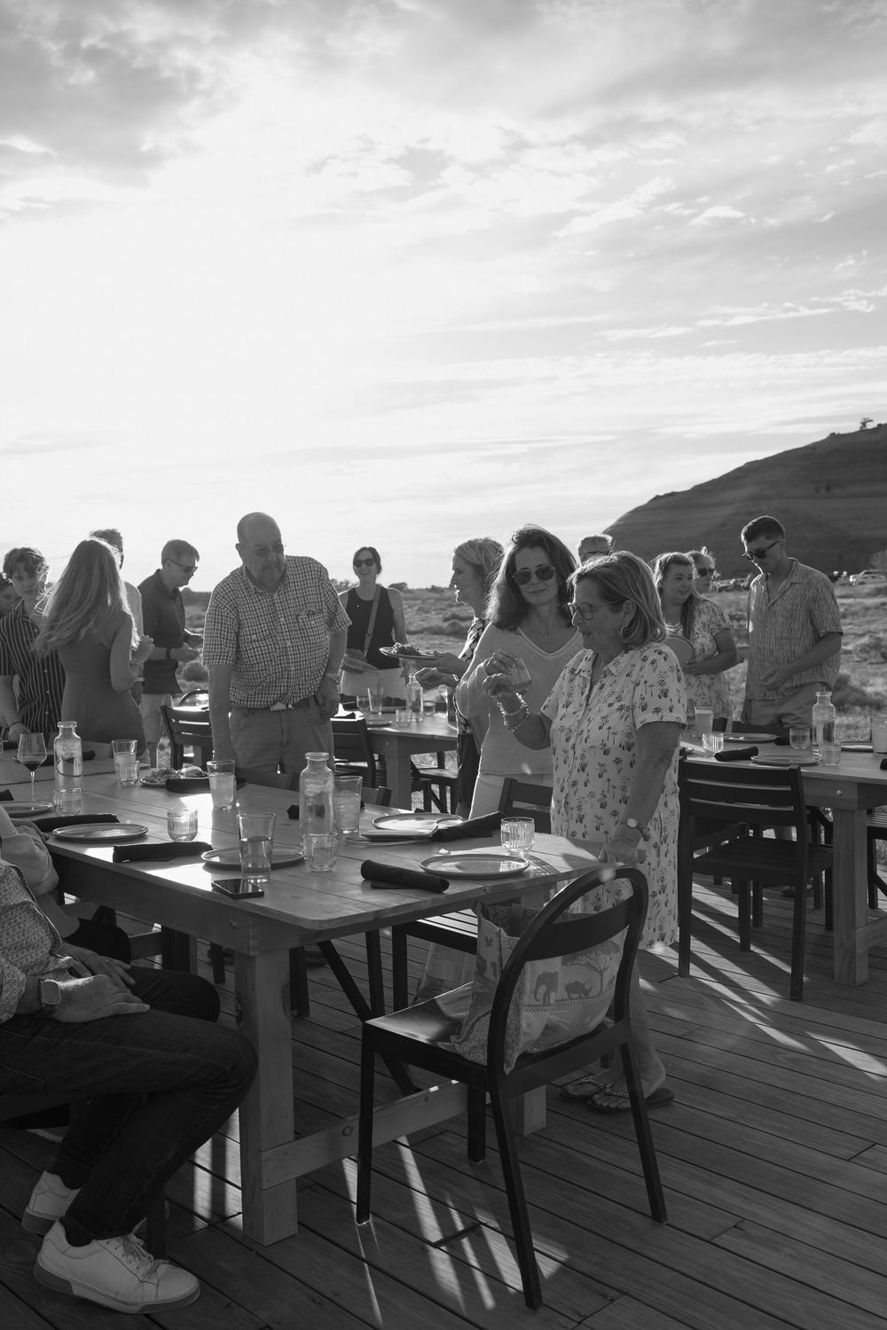 Black and white photo of guests socializing at an outdoor dining event on a wooden deck at sunset.