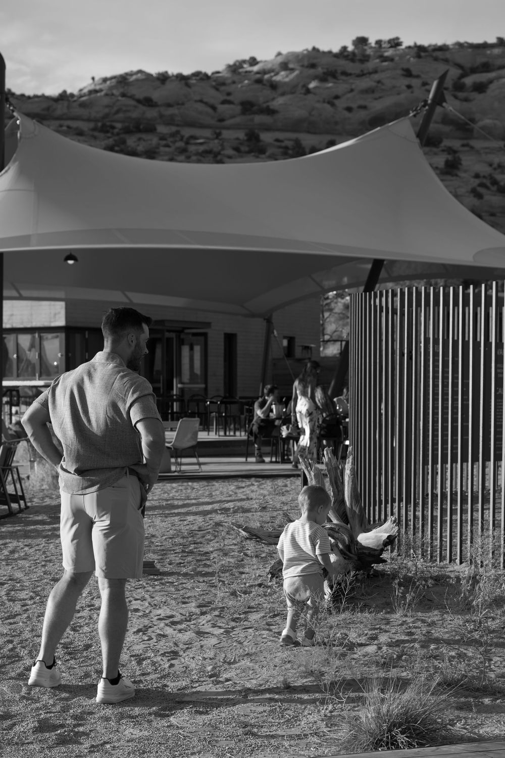 Black and white photo of a man and toddler walking toward an outdoor desert restaurant with a white tensile canopy.