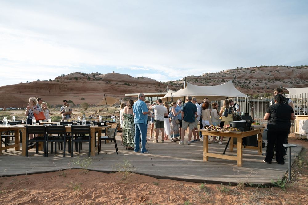 Outdoor gathering on a wooden deck with red rock desert landscape backdrop, guests mingling under white canopy tents.