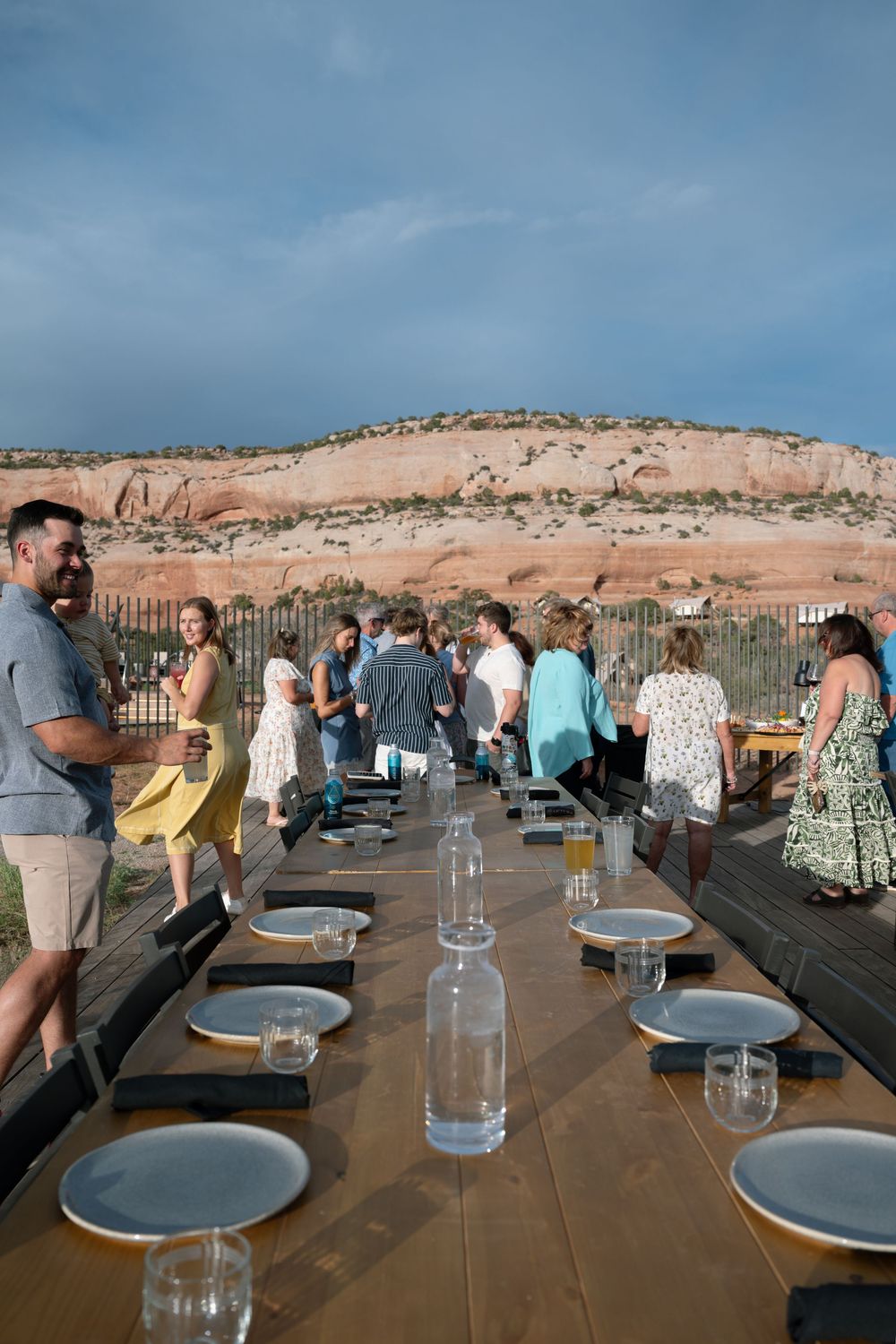 Outdoor dinner party on wooden deck with red rock canyon backdrop, guests mingling at long table set with plates and glasses.