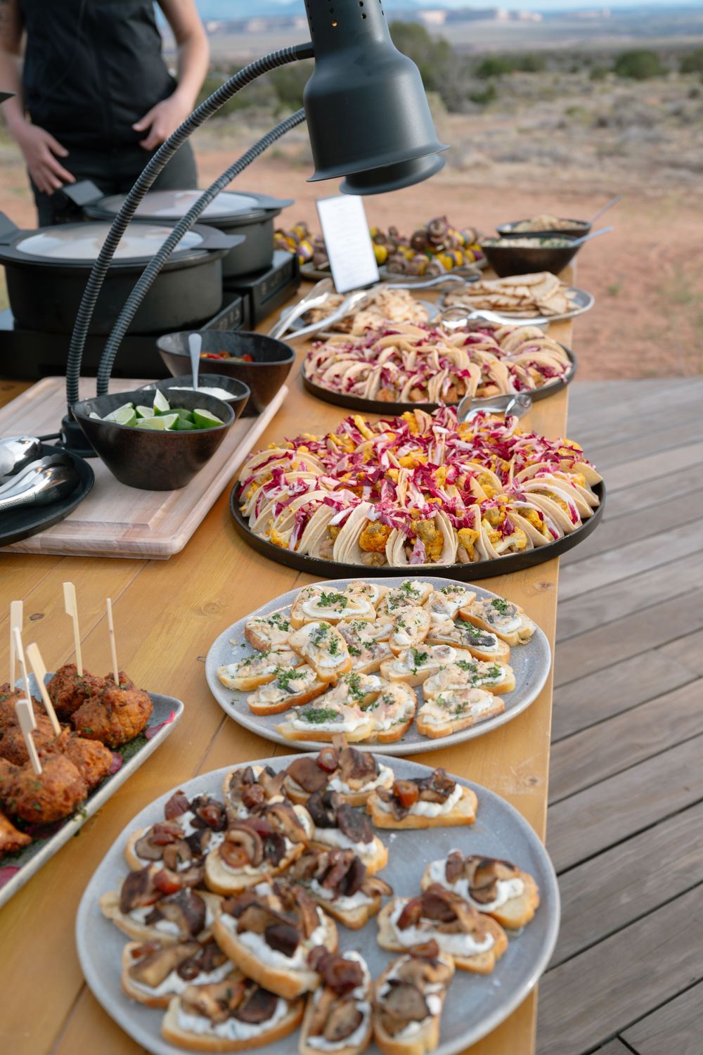 Outdoor catered buffet table with assorted appetizers, bruschetta, and finger foods displayed on plates in a desert landscape.