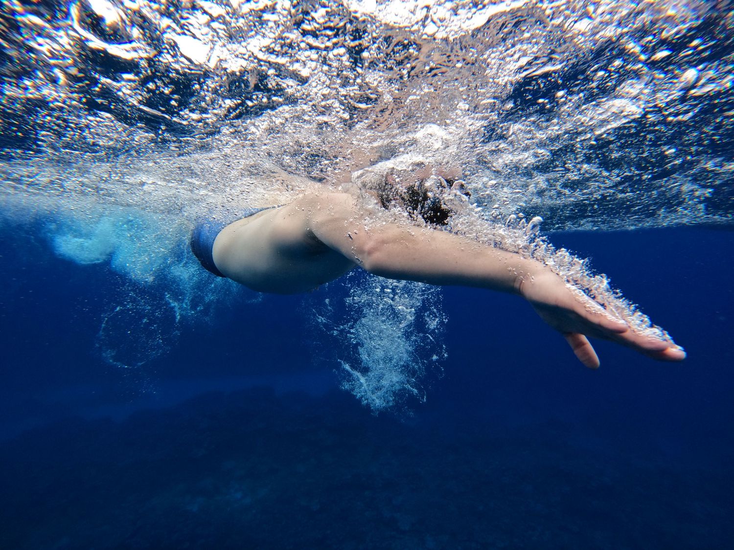 Underwater view of someone diving into deep blue ocean waters creating dramatic splash patterns.