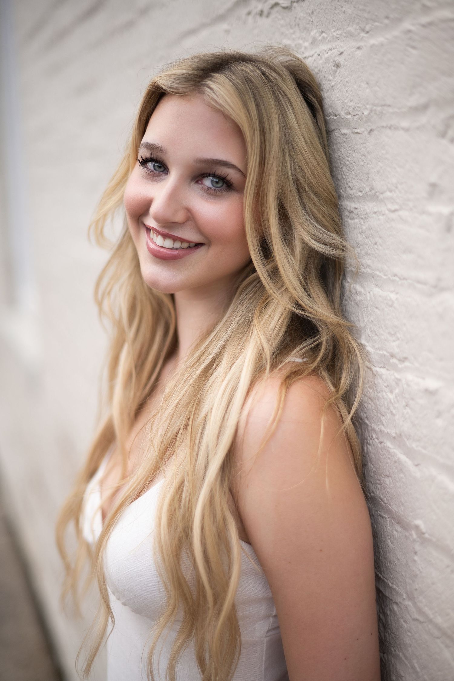Young blonde woman with long wavy hair smiling against a white brick wall, wearing a white sleeveless top.