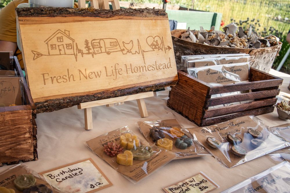 Rustic wooden sign for Fresh New Life Homestead displayed at a market booth with handmade candles and soaps on a linen-covered table.