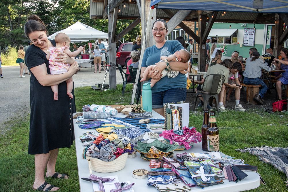 Two women holding babies browse handmade goods at an outdoor craft fair vendor table on a sunny day.