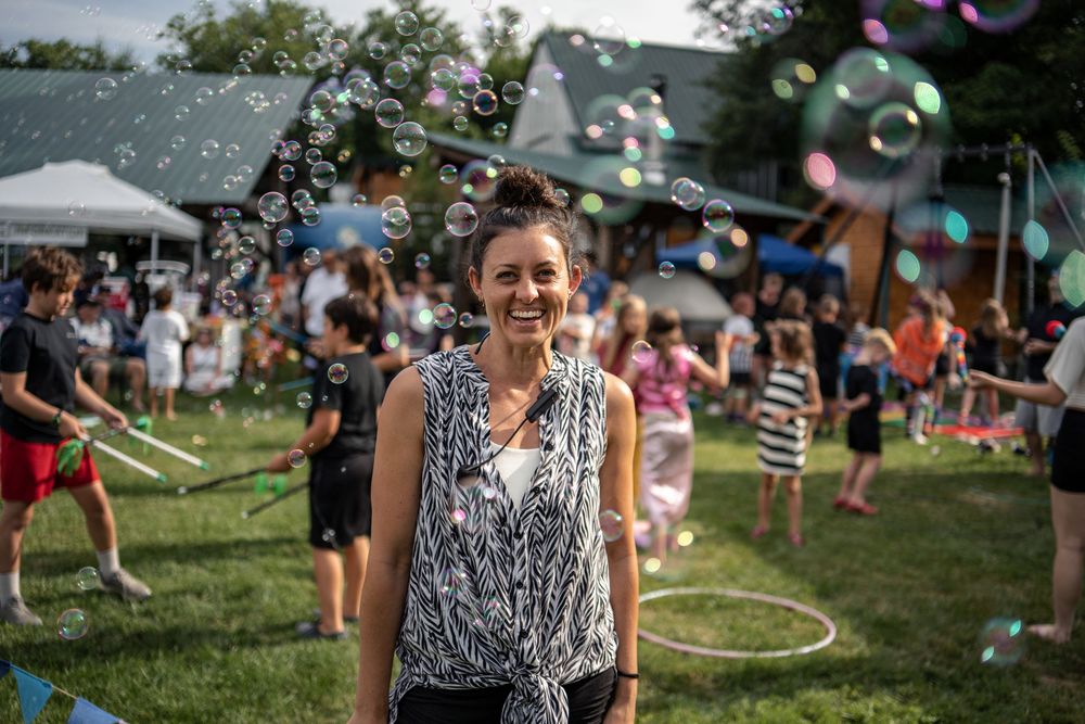 Woman smiling outdoors at a festive park event surrounded by colorful floating bubbles and people playing on green grass.