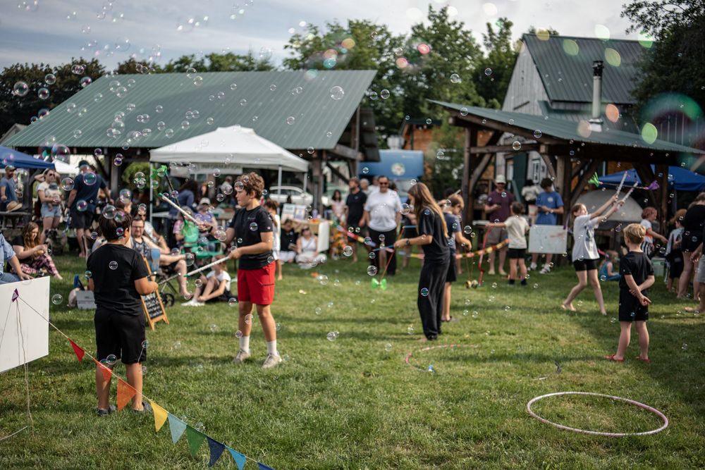 People enjoying an outdoor festival with bubble performers on green grass, tents and crowds in the background.