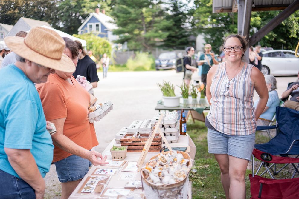 People browsing outdoor market stall with baked goods, smiling vendor in plaid shirt serving customers at summer community event.