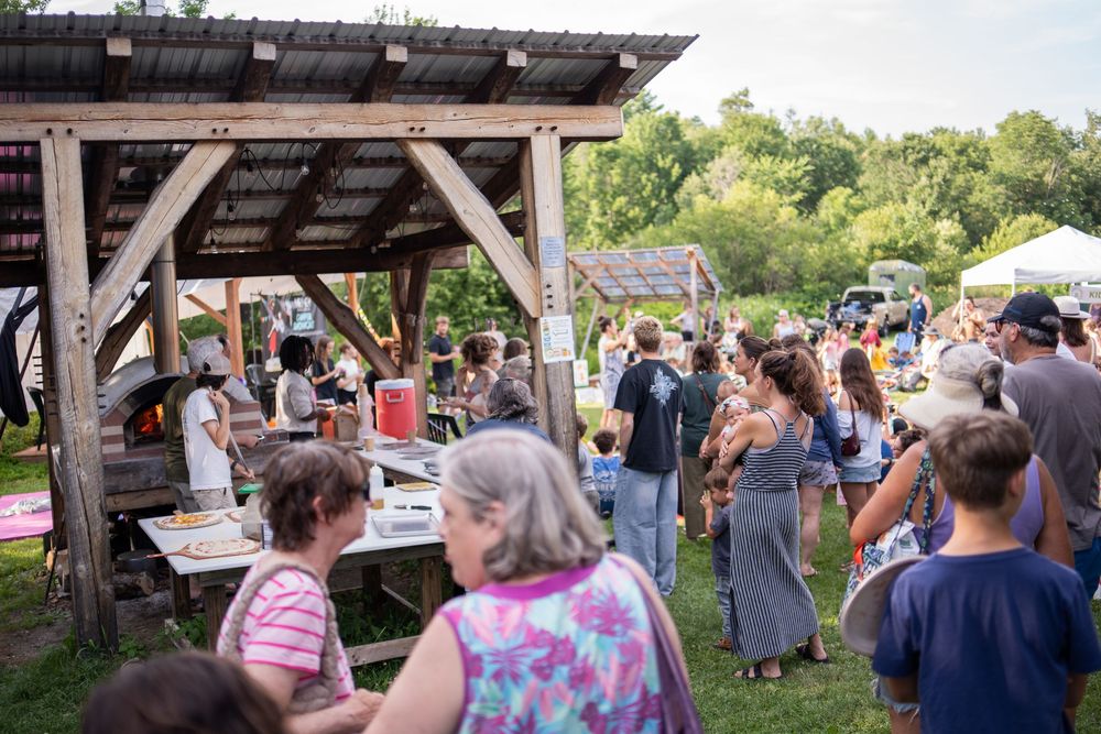 Crowd of people gathering at an outdoor farm event under a rustic wooden pavilion surrounded by lush greenery.