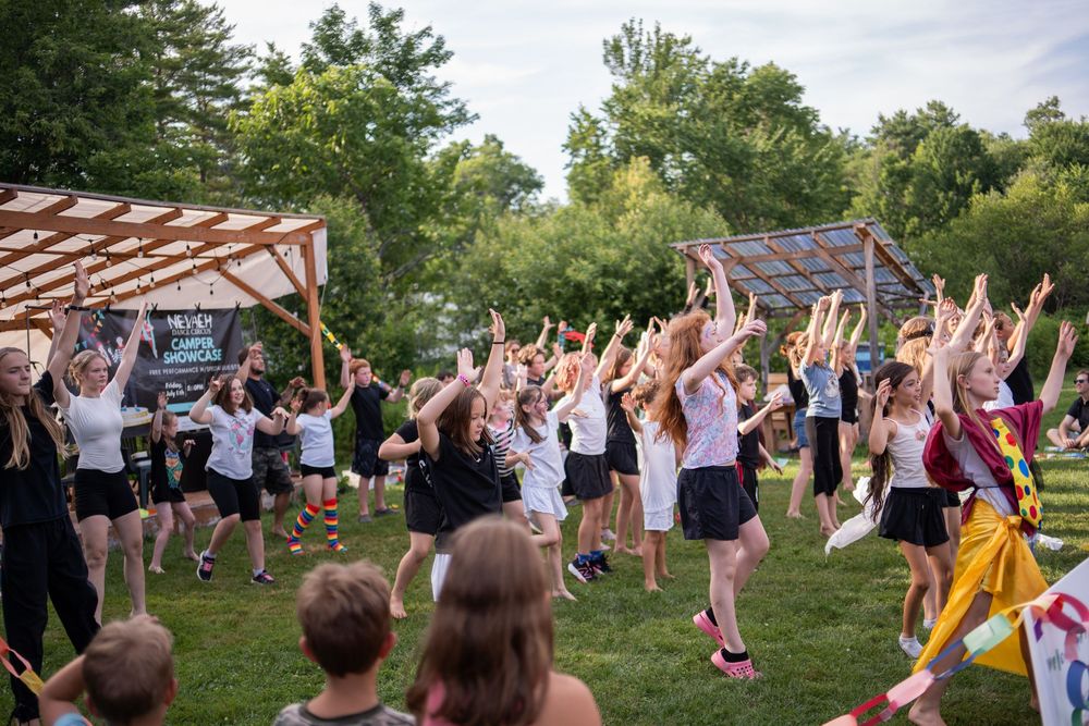 Large group of children and adults dancing outdoors at a summer camp event on a green lawn surrounded by trees.