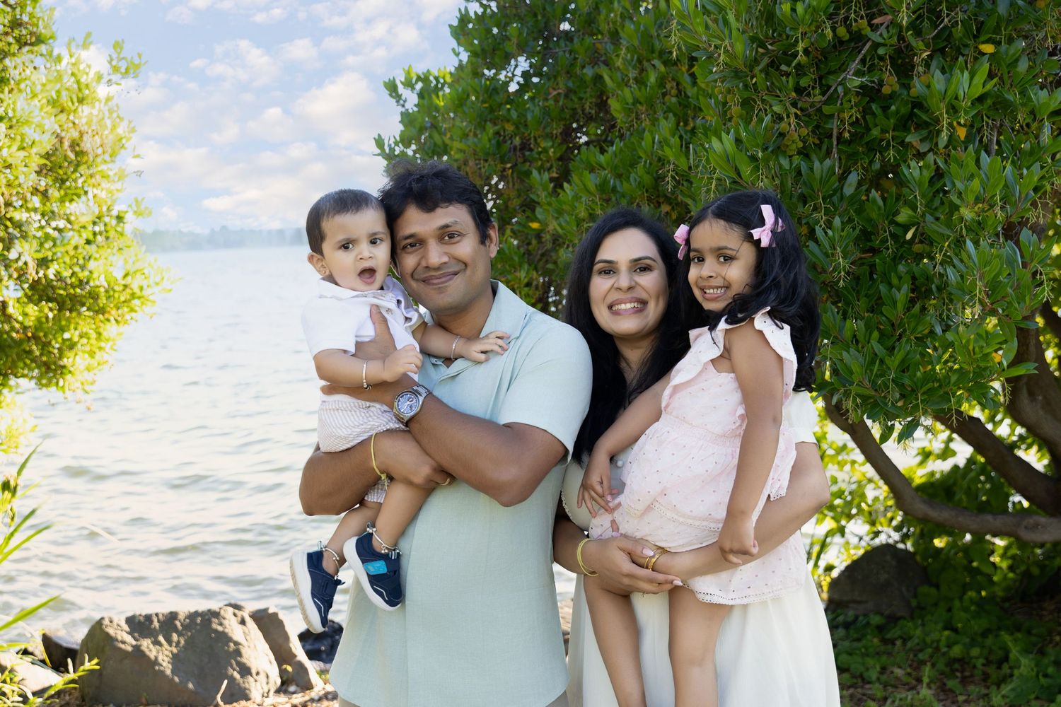 Happy South Asian family of four posing outdoors near waterfront, parents holding two young children among green trees.